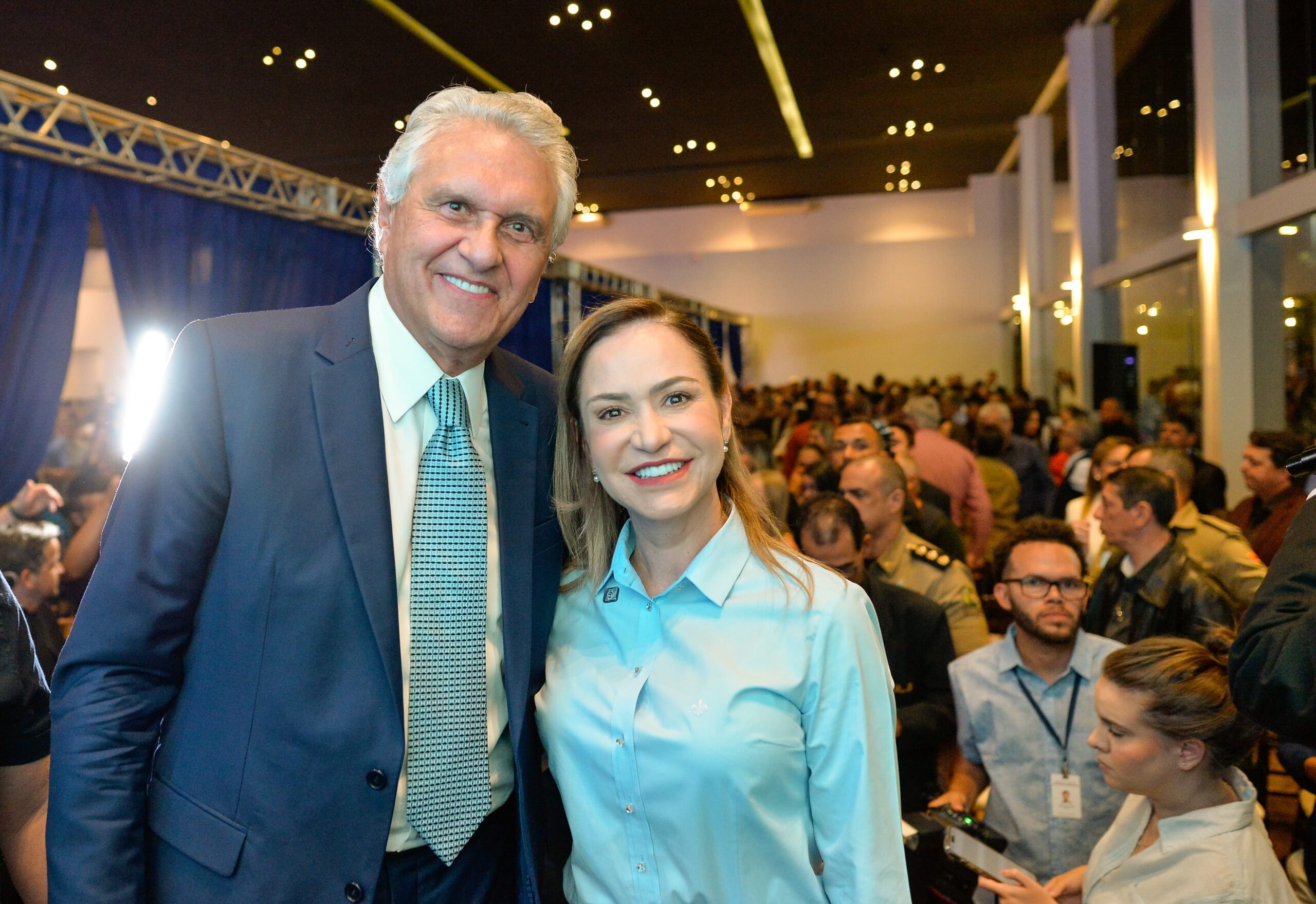 Sala de estar com móveis claros e buffet bege encostado nas costas do sofá com vasos em cima e itens de decoração, com uma parede de vidro e as demais em madeira, e a mesa da sala de jantar é bege escura com duas cadeiras estofadas em roxo e uma poltrona branca